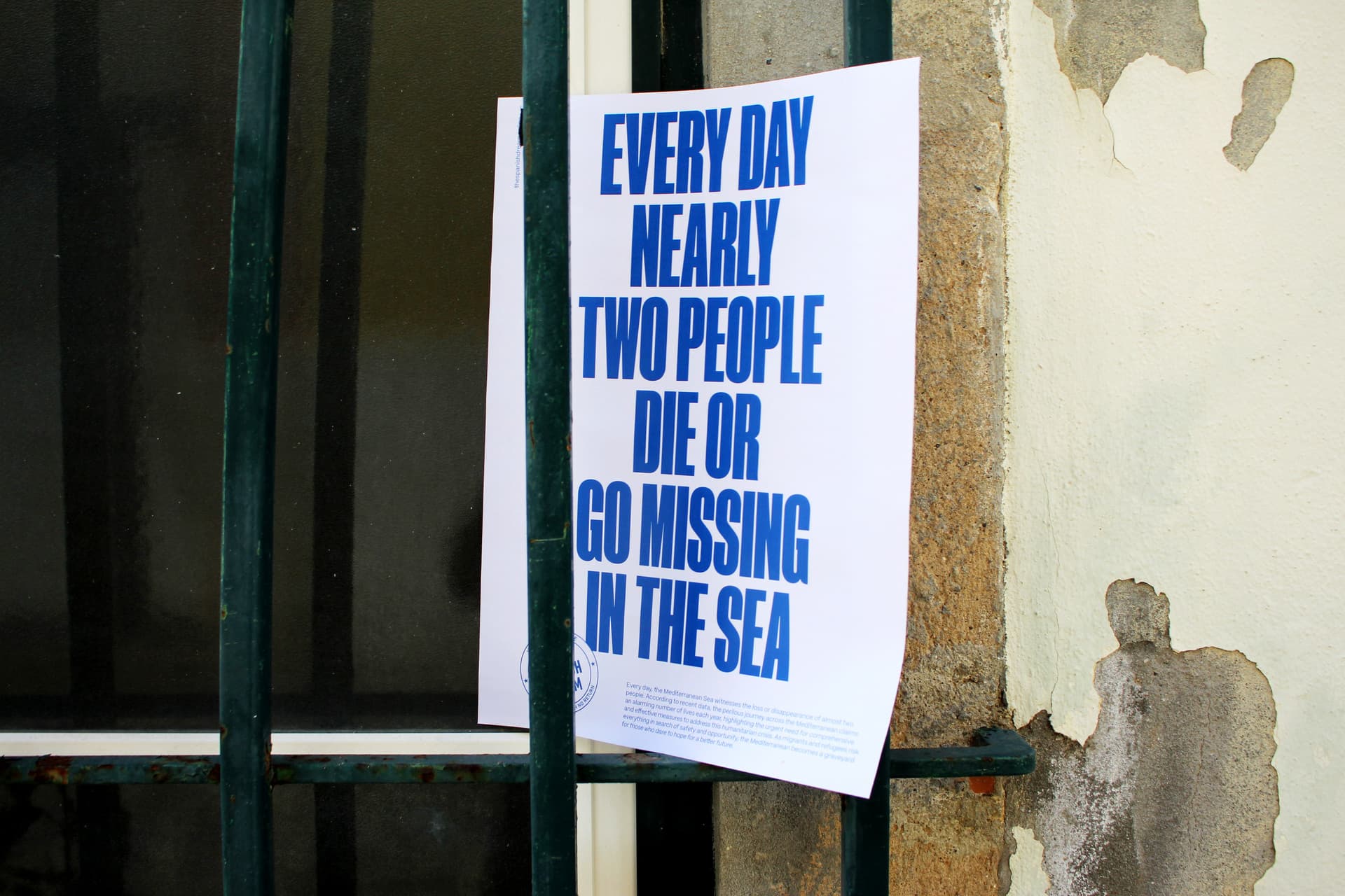 Cartel blanco con texto grande en azul que dice 'Every day nearly two people die or go missing in the sea', parte de la campaña 'The Spanish Dream', pegado en una pared exterior desgastada.