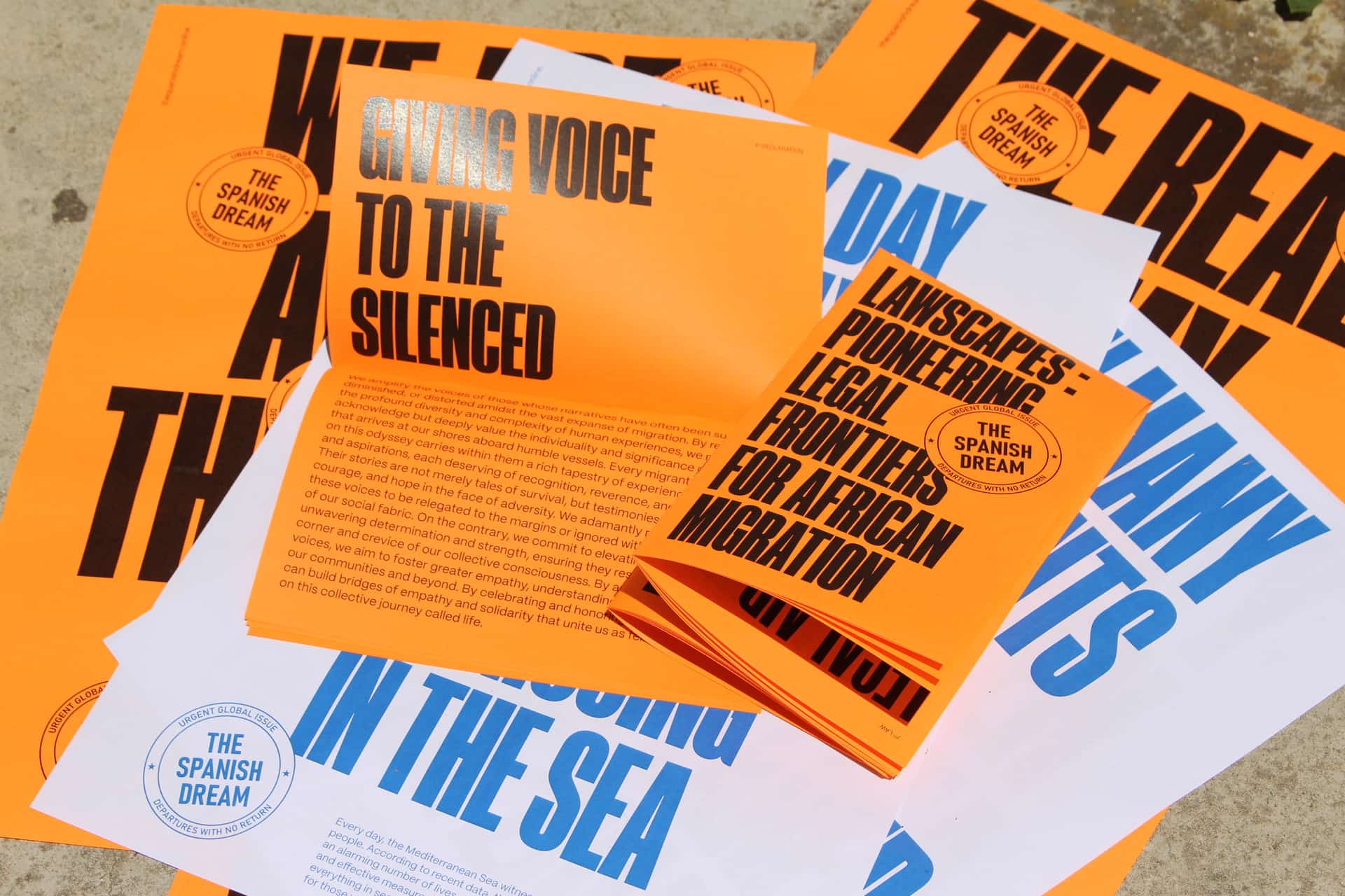 Composición de diversos materiales impresos del proyecto 'The Spanish Dream', incluyendo publicaciones naranjas con el titular 'Giving Voice to the Silenced' y un folleto blanco y azul sobre fronteras legales para la