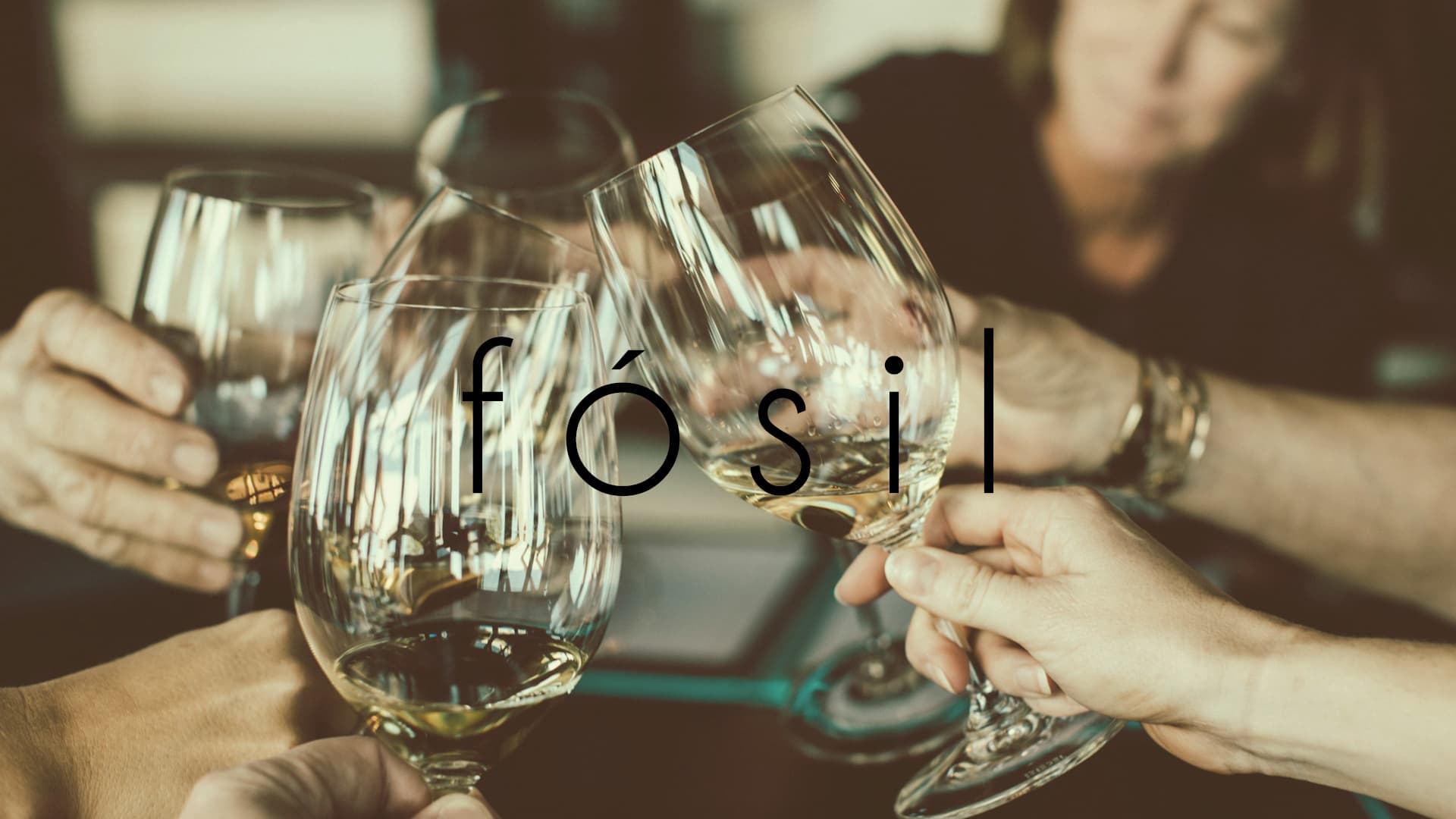 Fotografía de ambiente con varias copas de vino blanco en un brindis, el logotipo 'fósil' superpuesto elegantemente, sugiriendo un momento de celebración.