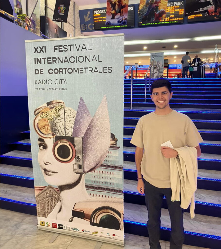 Joan Sanchis sonriente junto a un roll-up publicitario del XXI Festival Internacional de Cortometrajes Radio City, con diseño de collage surrealista, en la entrada de un cine.