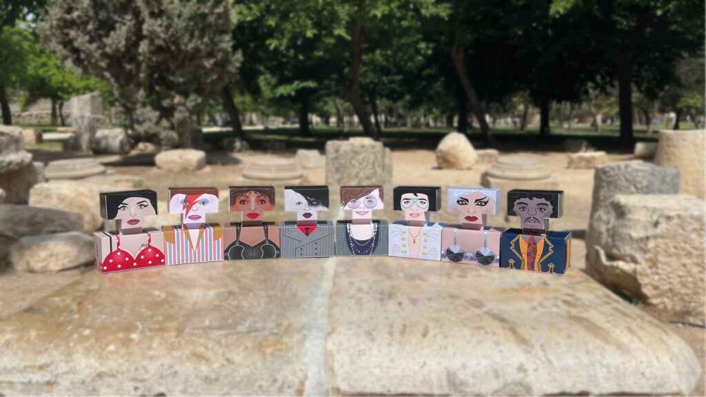 Colección de figuras de cartón o madera con forma de bloque, representando a iconos como Amy Winehouse, David Bowie, y Nina Hagen, dispuestas al aire libre sobre piedra