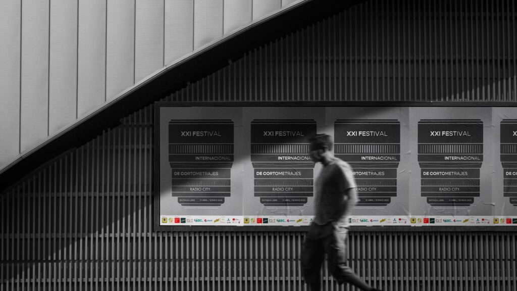 Serie de carteles del XXI Festival Internacional de Cortometrajes Radio City en una pared de listones, con una persona desenfocada caminando delante, mostrando la aplicación del diseño en un espacio público.