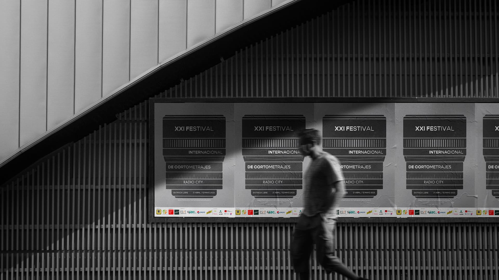 Serie de carteles del XXI Festival Internacional de Cortometrajes Radio City en una pared de listones, con una persona desenfocada caminando delante, mostrando la aplicación del diseño en un espacio público.
