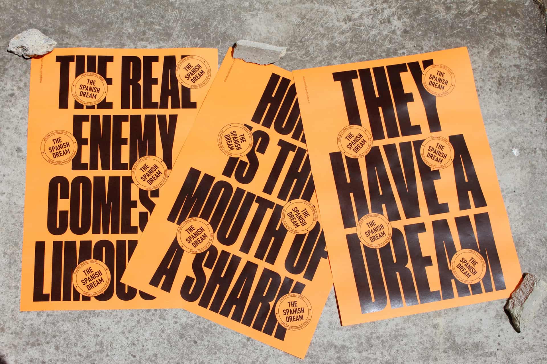 Tres carteles grandes de color naranja con tipografía negra del proyecto 'The Spanish Dream', con frases como 'The real enemy comes in a limousine' y 'They have a dream', extendidos en un suelo de cemento.