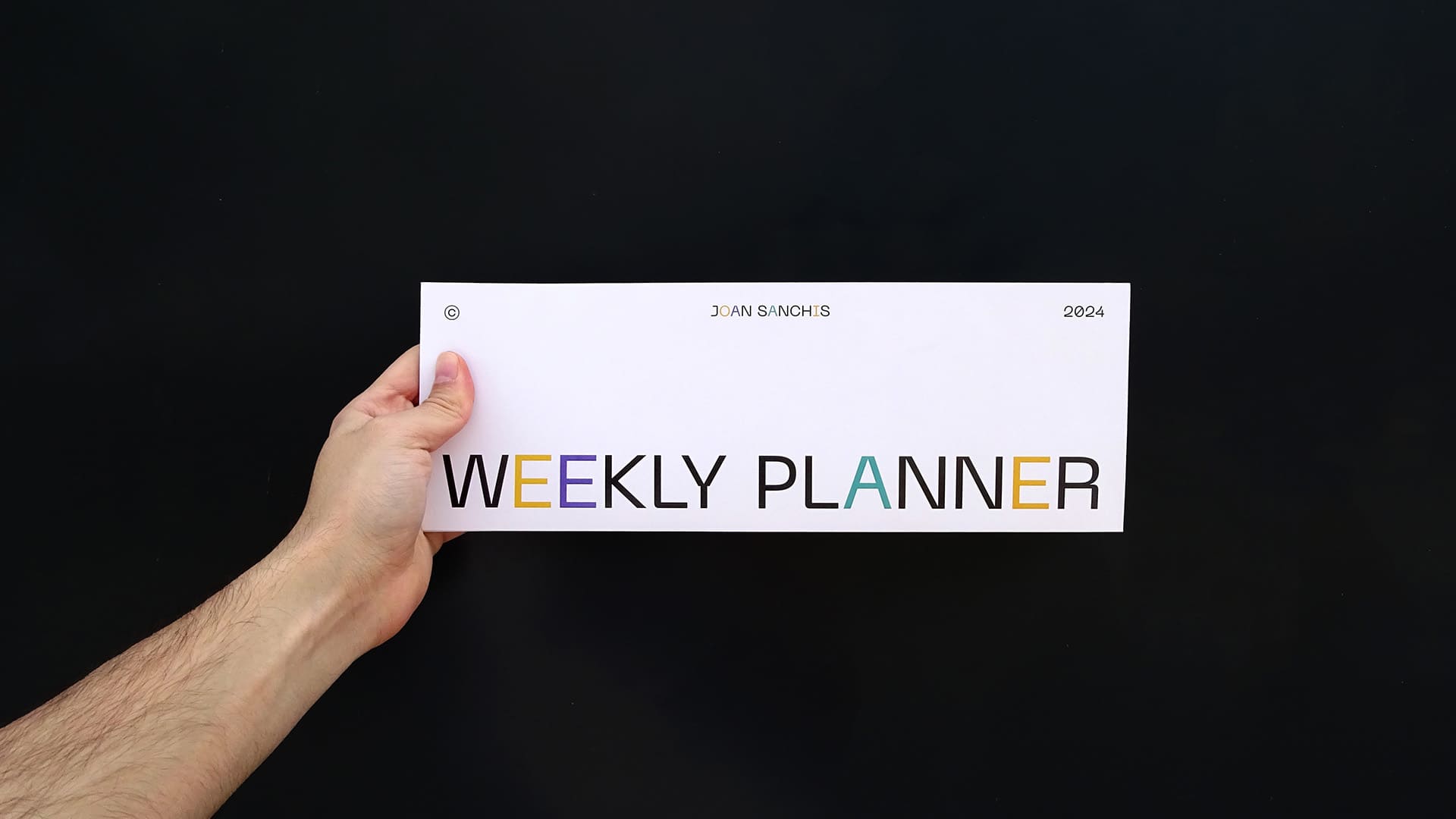 Mano sosteniendo la portada de un planificador semanal horizontal diseñado por Joan Sanchis, con el título 'WEEKLY PLANNER'