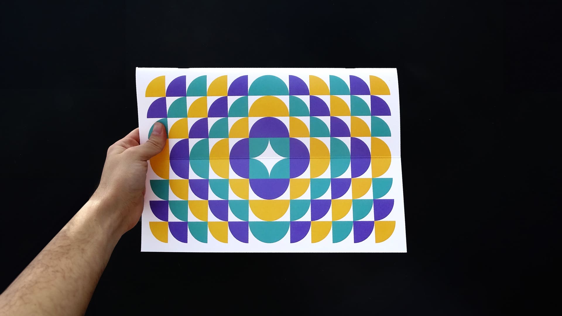 Mano sosteniendo una hoja con un patrón geométrico abstracto en colores morado, turquesa y amarillo, parte del diseño del planificador, sobre un fondo negro