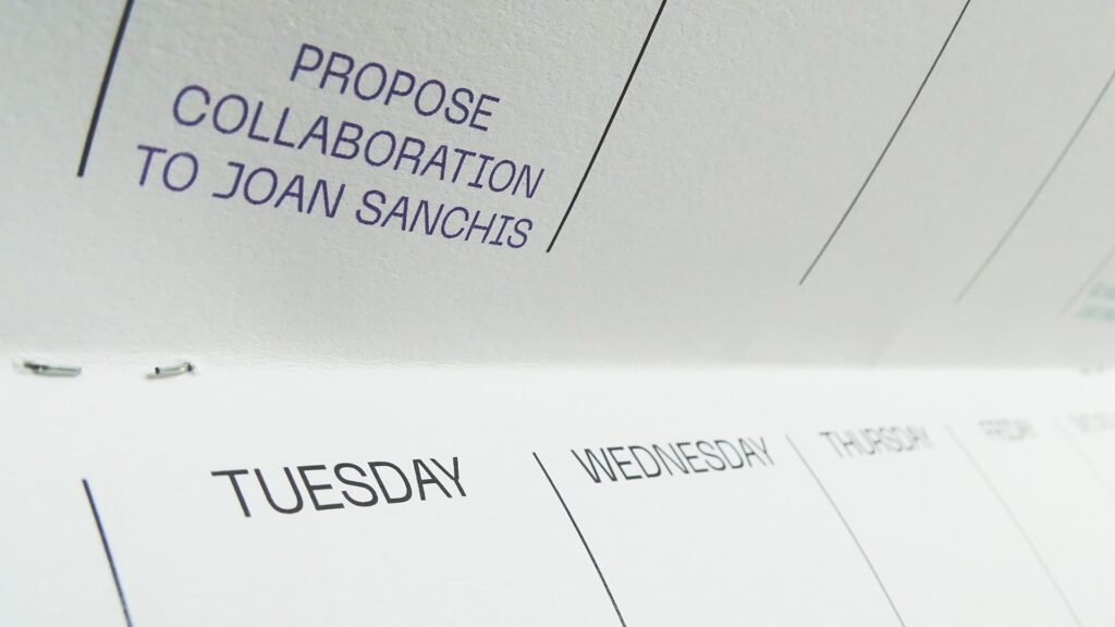 Primer plano de una casilla del planificador semanal con el texto 'PROPOSE COLLABORATION TO JOAN SANCHIS' en tinta morada, y los días 'TUESDAY' y 'WEDNESDAY' visibles